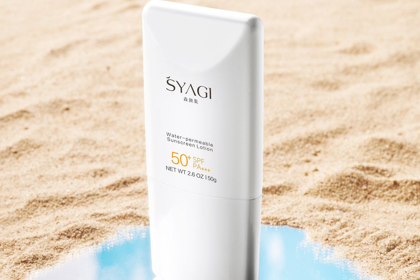 A sunscreen bottle standing on sand beside a mirror reflecting blue sky at the beach.