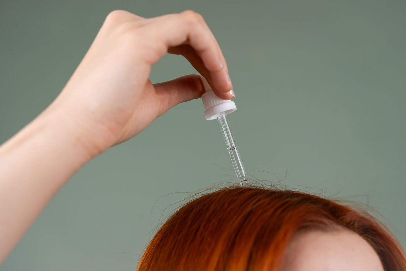 Close-up of a hand applying hair serum to red hair using a dropper.